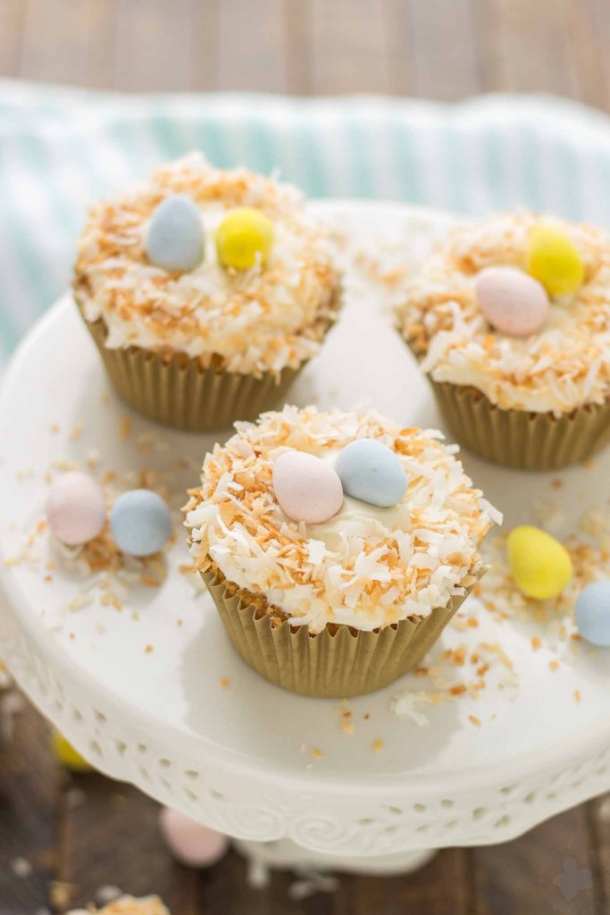 3 coconut cupcakes garnishes with Easter eggs sitting on a white cake stand.