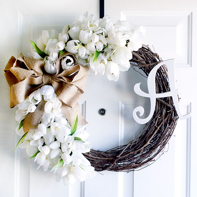A homemade wreath with white tulips and a gold bow hanging on a white door.